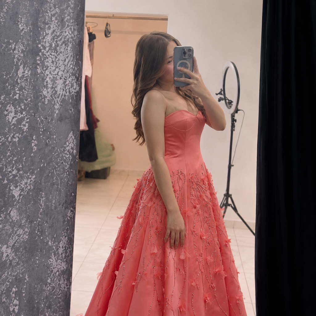 A gorgeous woman takes a mirror selfie in a fitting room wearing a coral pink strapless ball gown with a structured bodice and a voluminous skirt featuring shiny detailed beadwork and 3D floral applique.