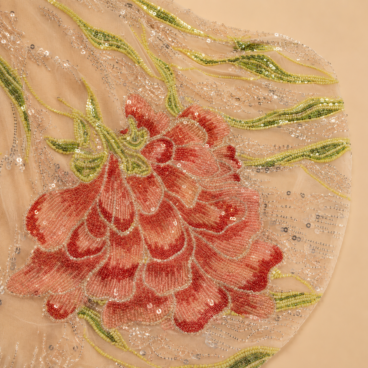 Close-up of a sheer beige embroidered embellished fabric from the train of a luxury evening gown featuring a large pinkish-red flower with green leaves and silver sequins. 