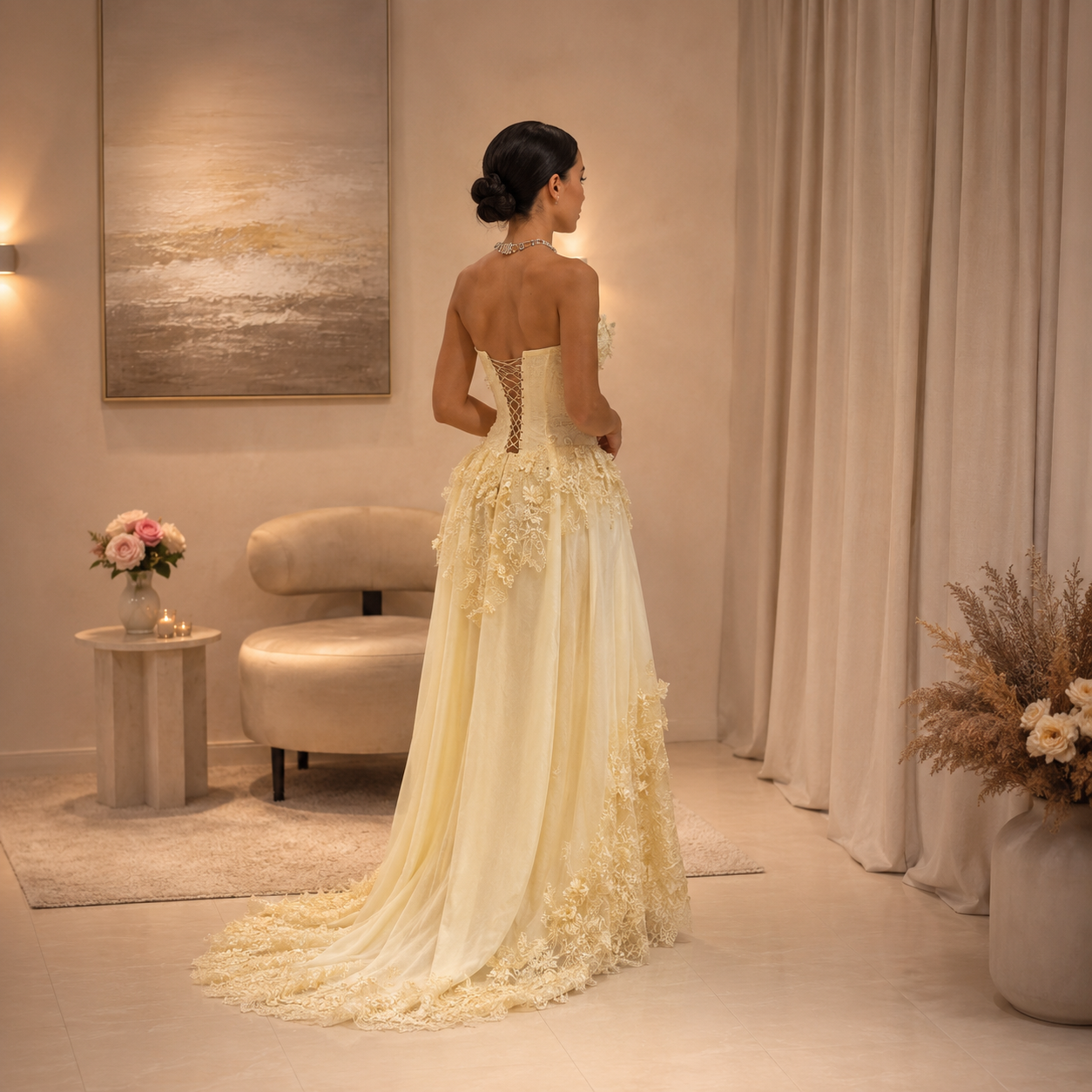 A woman stands with her back to the camera wearing a strapless pale yellow luxury evening gown with 3D floral appliqués and a long train in a softly lit indoor room with beige walls, curtains, and a small upholstered chair.