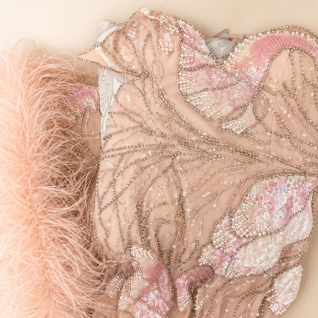 Close up of a luxury blush pink evening gown, with soft pink feather accessory and detailed shimmering crystal beadwork embellishments all over the stunning dress