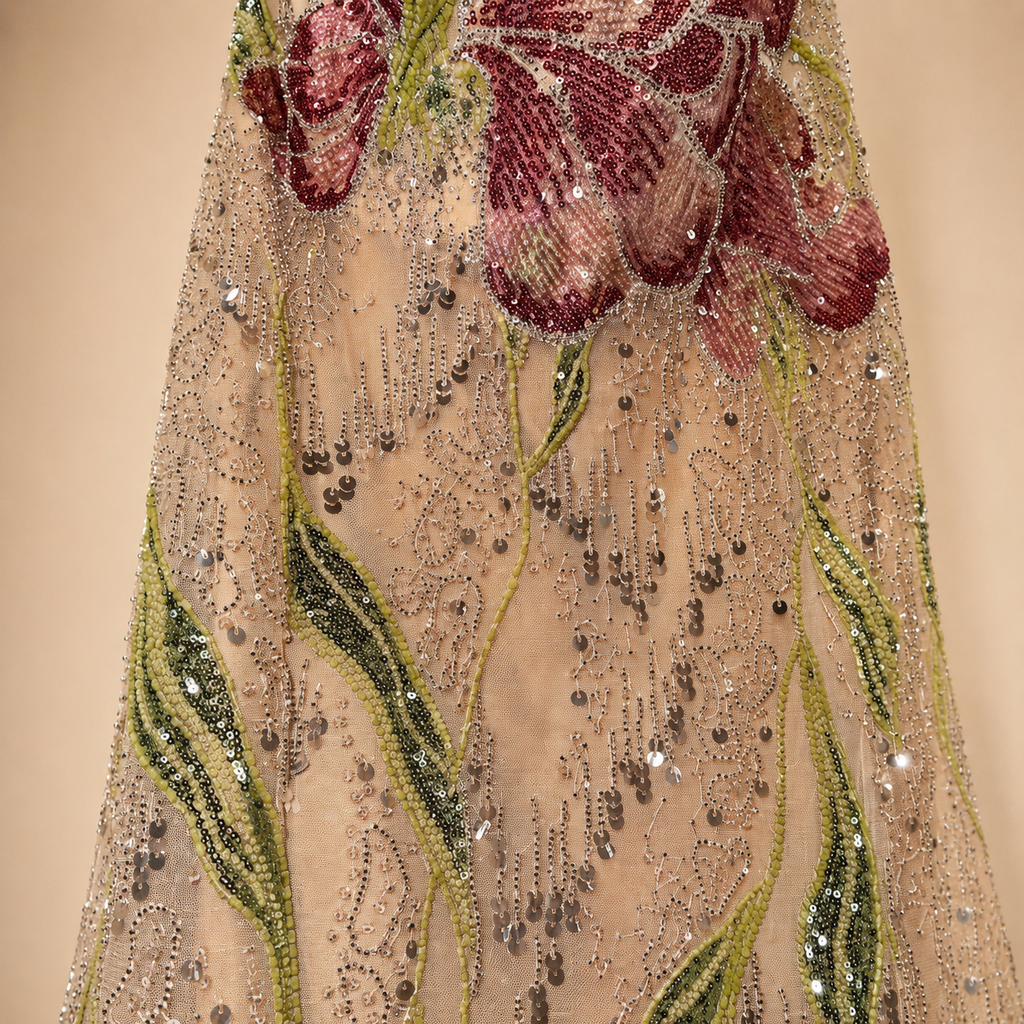 Close-up of a heavily embellished sheer beige champagne fabric of a luxury-fashion evening gown dress with large burgundy floral motifs and green leaf patterns covered in silver and metallic beadwork.