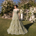 Beautiful lady in a luxurious sage green princess ballgown dress, the gown has cascading petal details and is covered with shimmering embellishments, detailed beadwork. There are Spaghetti straps and delicate off shoulder accent sleeves. She is in a picturesque outdoor setting with flowers in the background