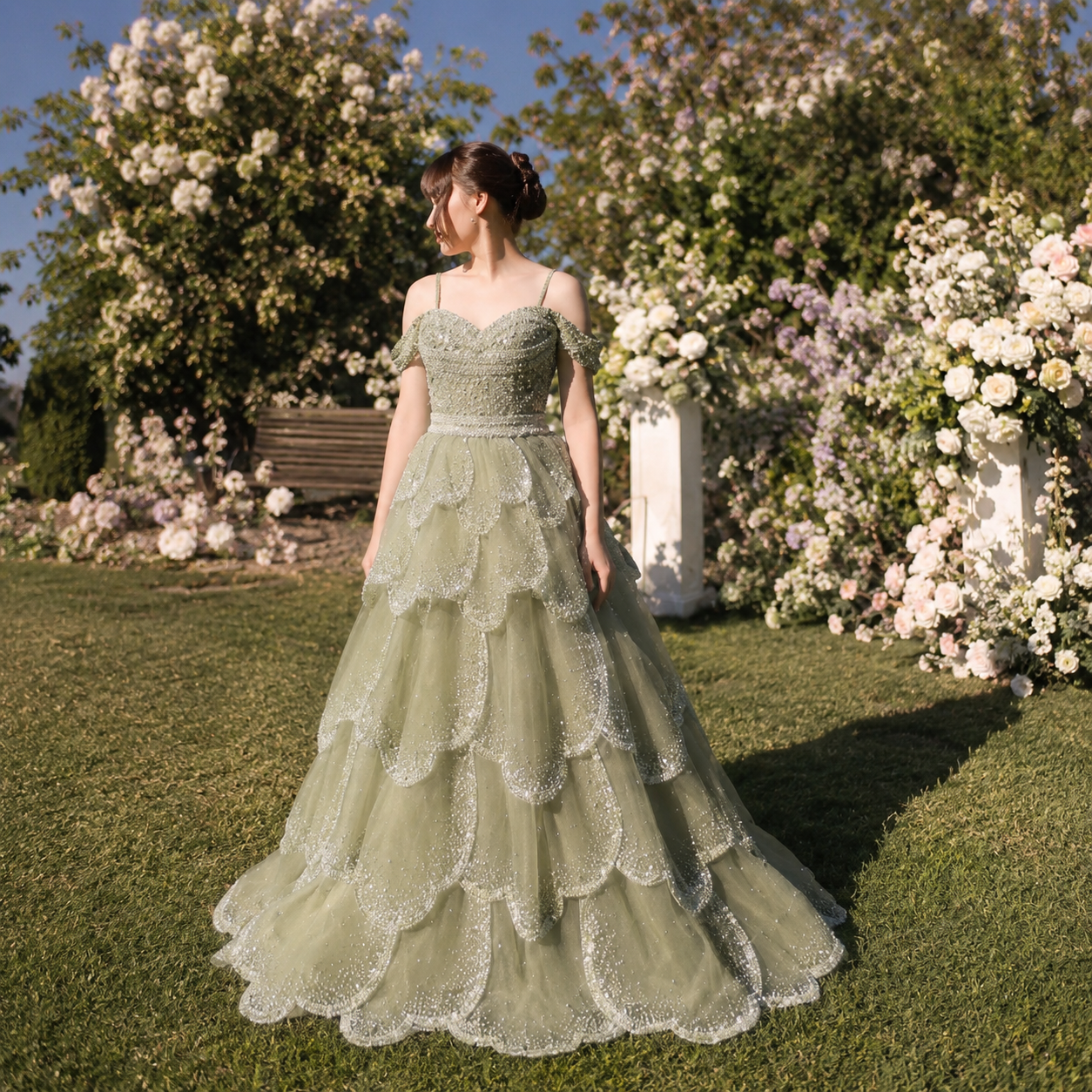 Beautiful lady in a luxurious sage green princess ballgown dress, the gown has cascading petal details and is covered with shimmering embellishments, detailed beadwork. There are Spaghetti straps and delicate off shoulder accent sleeves. She is in a picturesque outdoor setting with flowers in the background
