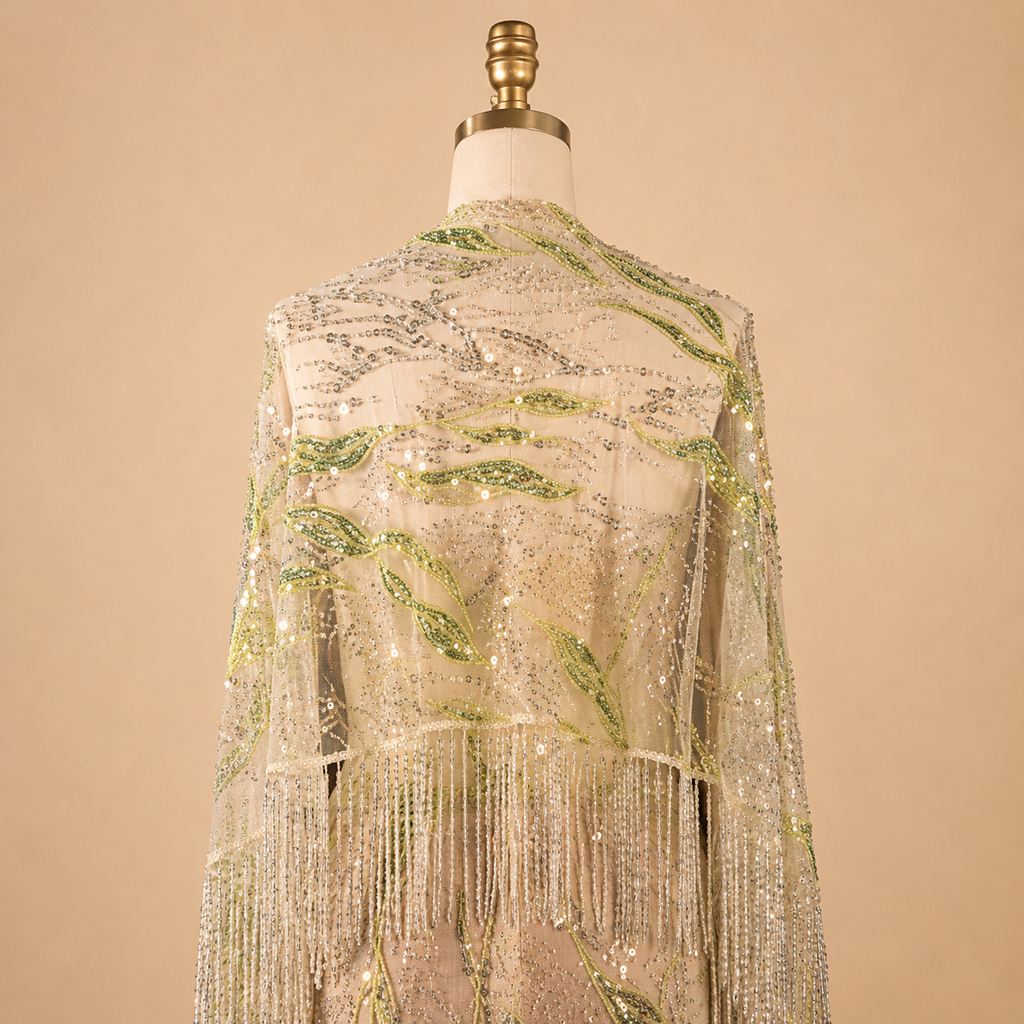 A luxury shimmering beaded fringe shawl or wrap displayed on a dress form mannequin. The garment features intricate silver sequins and beadwork with embroidered green leaf patterns, set against a solid beige background.