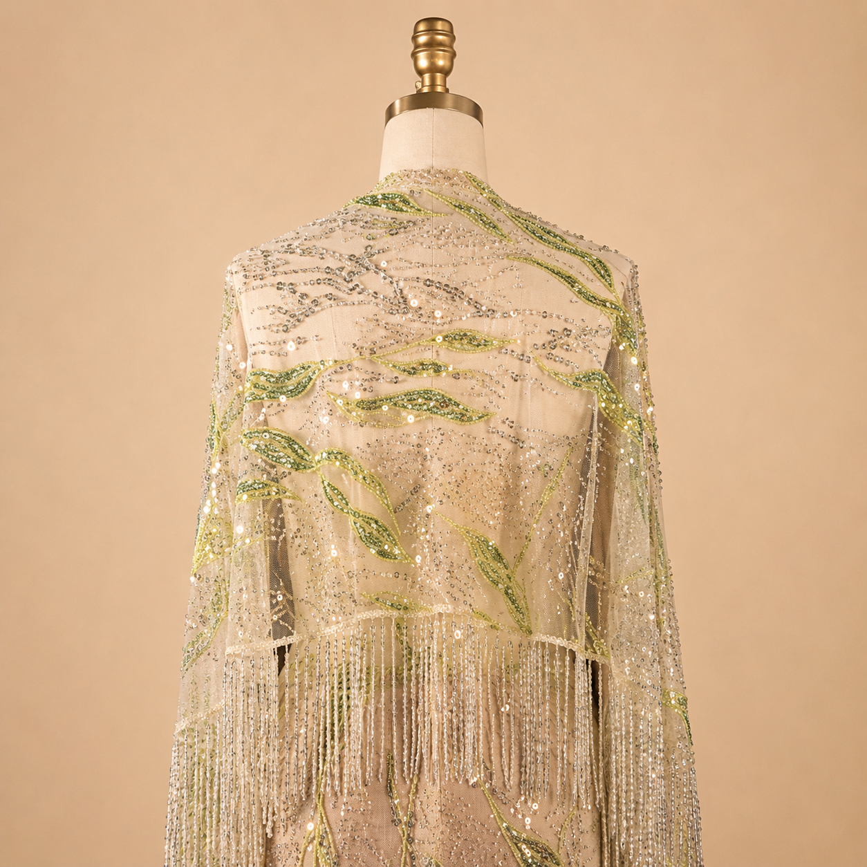 A luxury shimmering beaded fringe shawl or wrap displayed on a dress form mannequin. The garment features intricate silver sequins and beadwork with embroidered green leaf patterns, set against a solid beige background.