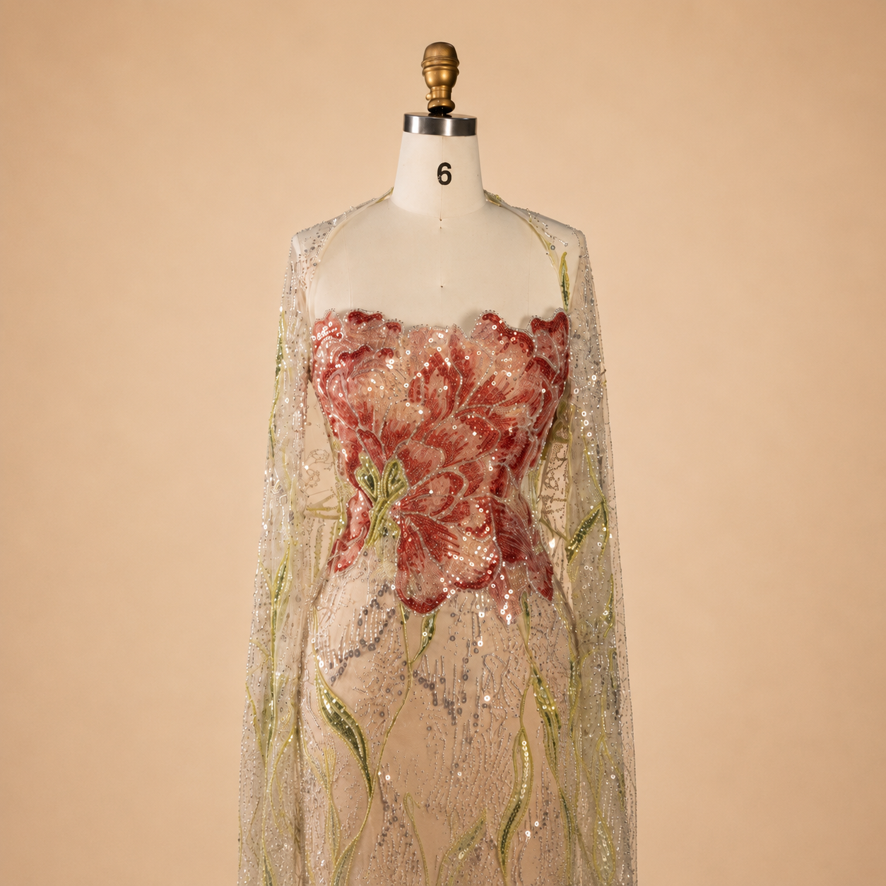 A sheer beige champagne sequined luxury gown displayed on a white mannequin against a warm beige background. The bodice features a prominent shimmering red floral applique with green embroidered vine details.