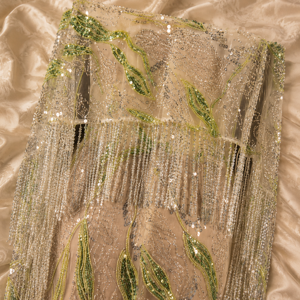 A close-up of a champagne-colored sequined fabric of a luxury dress with green embroidered leaf motifs and cascading bead fringe, laid flat on a matching satin fabric background.