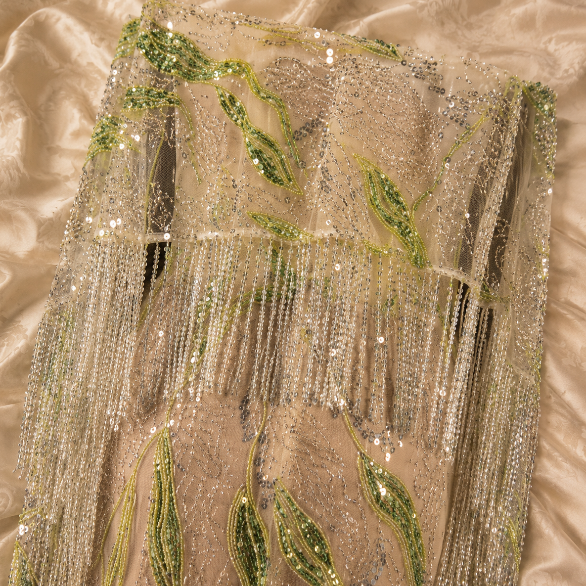 A close-up of a champagne-colored sequined fabric of a luxury dress with green embroidered leaf motifs and cascading bead fringe, laid flat on a matching satin fabric background.