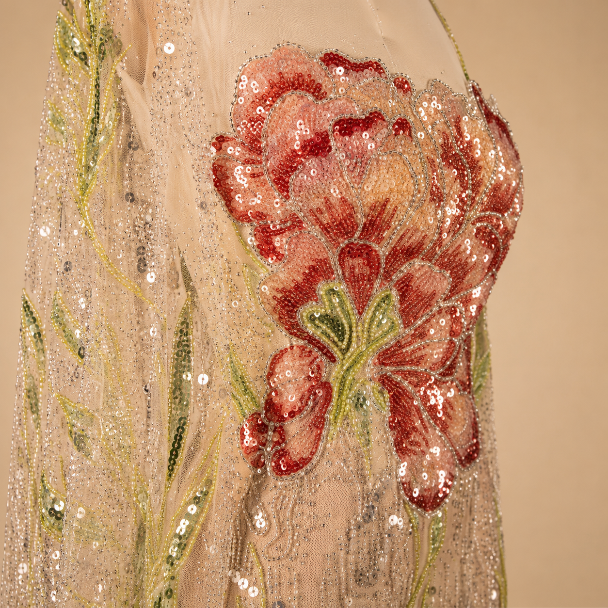 Close-up of a shiny champagne-nude sequin and beaded embellished garment featuring a large red and pink floral applique with green sequin vine details on a warm beige background.