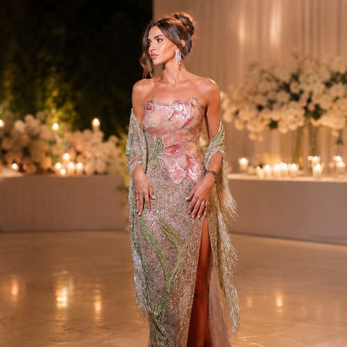 A woman models a luxury strapless embellished shimmering gown with a sheer draped shawl in a warm, elegant indoor setting with floral arrangements and candles in the background.