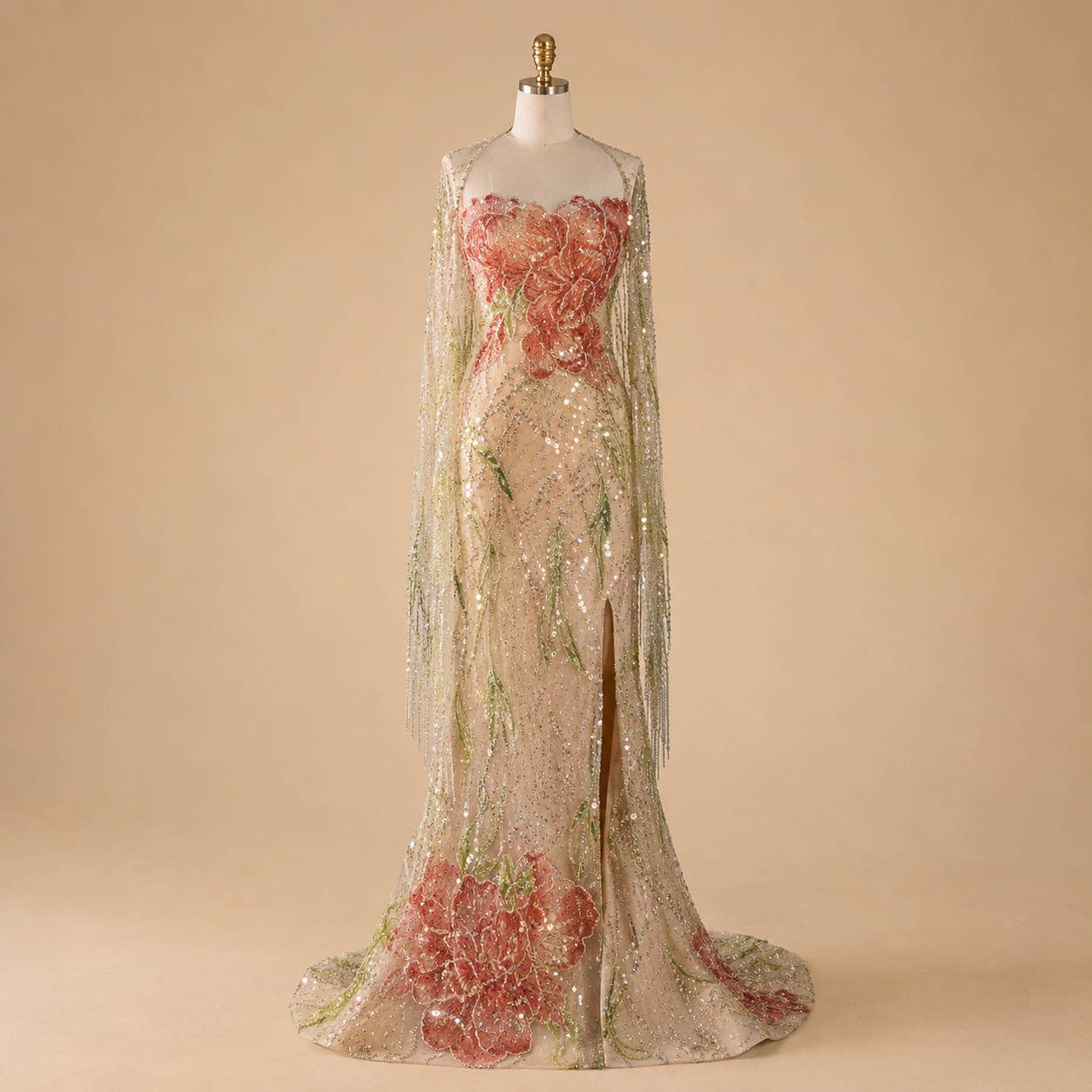 A luxury formal floor-length evening gown displayed on a mannequin against a warm beige studio background. The dress has a sheer nude-beige champagne base covered in shimmering sequins and decorated with large pink floral appliqués and green embroidered vine details.