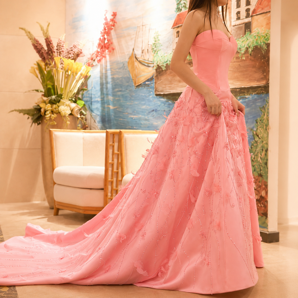 A woman wearing a strapless pink ball gown with a long train and floral embroidery, standing in an indoor setting with a painting and a chair in the background.